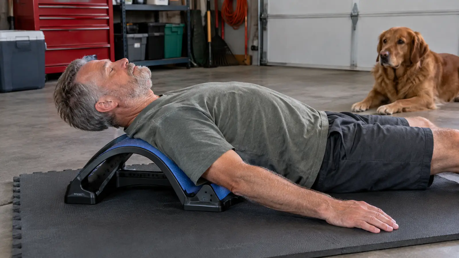 A person lying on a back stretcher on a foam mat in a garage while a dog watches from a few feet away
