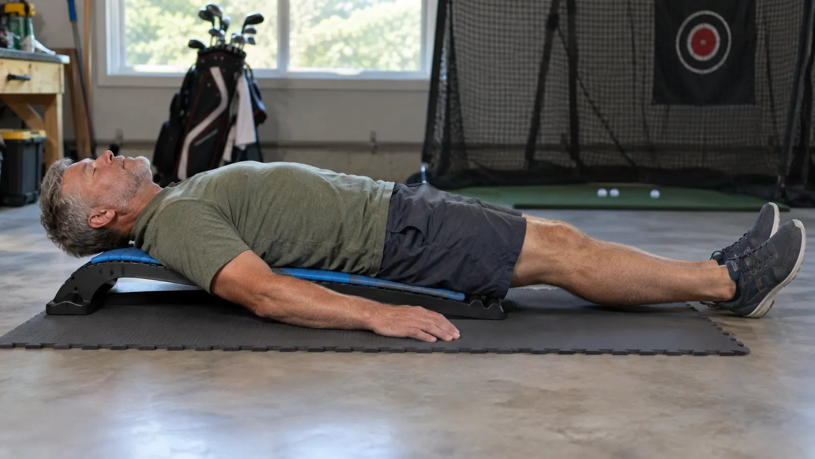 A person lying on a back stretcher on a foam mat in a garage, relaxed with arms at their sides