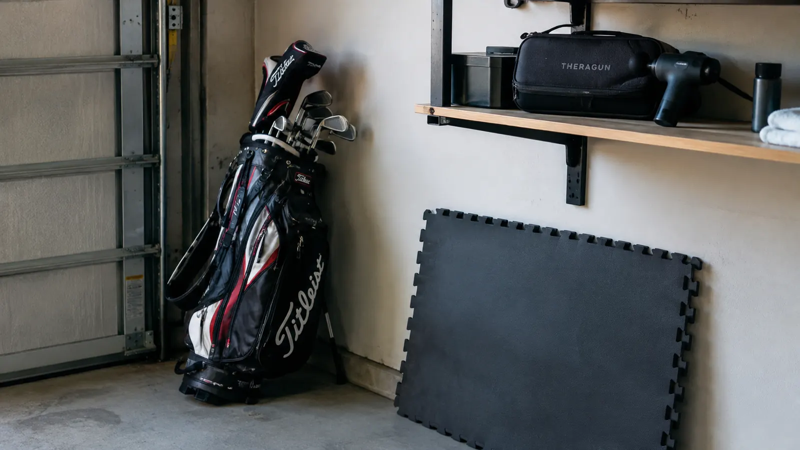 A golf bag and foam mat in a garage with a massage gun case on a shelf above